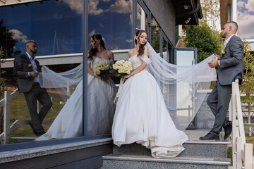 Groom assisting bride in elegant wedding attire outside a modern venue in daylight