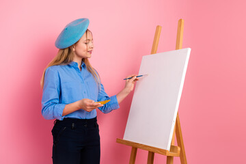 Young girl wearing a blue beret and shirt painting on a plain canvas in front of a pink background