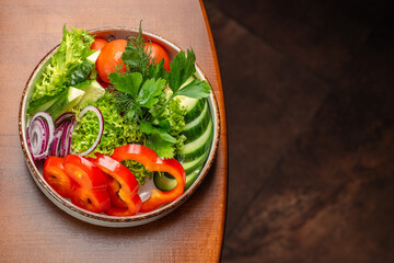 A fresh and colorful vegetable salad featuring sliced cucumbers, red bell peppers, tomatoes, red onions, lettuce, and fragrant herbs on a rustic wooden table
