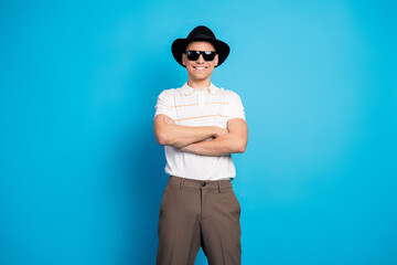 Confident young man with folded arms wearing sunglass and a hat, posing against a vibrant blue...
