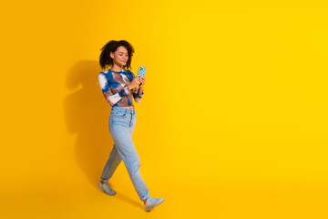 Stylish young woman with curly hair using smartphone against vibrant yellow background, showcasing...