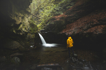 Hiker in a bright yellow rain jacket stands on damp stones beside a secluded forest waterfall,...