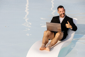 Man in blazer, swim trunks, and glasses sitting by the pool with a laptop, showing a thumbs-up...