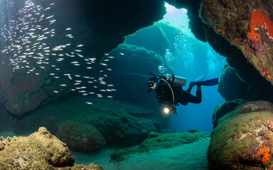 Scuba diver explores underwater cave system with school of fish and sunlight filtering down