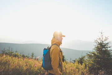 Man wearing a yellow jacket and a blue backpack stands in a field with a beautiful view of the...