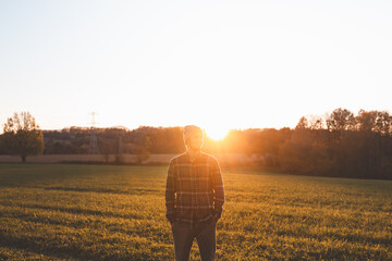 Young man in plaid shirt and beanie standing on green meadow at sunset. Golden sunlight, autumn...