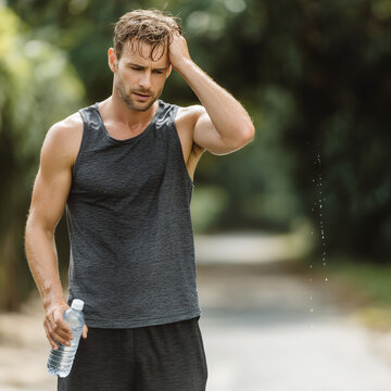 Athletic man holding water bottle, sweating after jog, greenery background