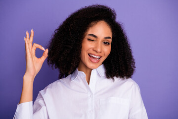 Attractive young woman in white shirt smiling with positive gesture and winking on a vibrant purple...