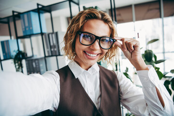 Smiling professional businesswoman in elegant attire taking a selfie in a modern office with a...
