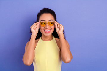 Young woman in yellow attire smiling beautifully while wearing trendy sunglass against a purple...