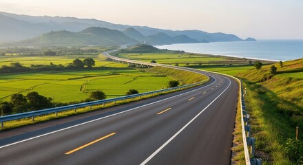 A winding asphalt road stretches along a coastline, with green fields and mountains in the background, under a clear sky.