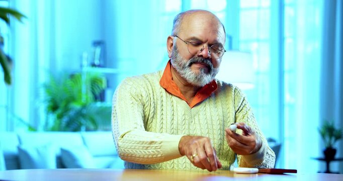 Indian senior man checking sugar level at home using handheld glucometer device, smiling happily after seeing normal reading, calling someone joyfully on smartphone to share positive health update