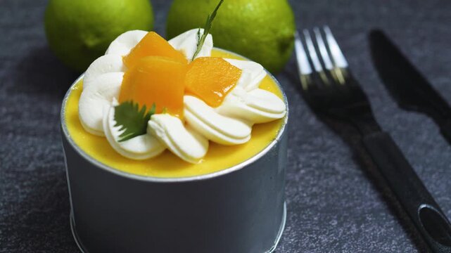 Delicate mango mousse cake Tin Can garnished with whipped cream, fresh peach slices, mint sprig, resting on dark textured background with limes, cutlery nearby