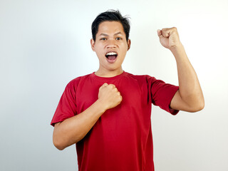 Excited Man Expresses Happiness and Victory Wearing a Red Shirt. Posing with his right fist clenched at his chest and his left hand raised upward.