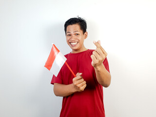 A cheerful Asian man in red shirt holds an Indonesian flag and waves it, making a love sign with...