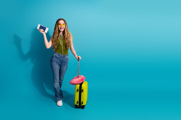 Smiling young woman in casual summer fashion holding passport with a suitcase against a blue...