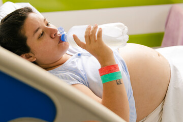 Pregnant woman with identification bracelets on her wrist drinking water from a bottle while lying in a hospital bed