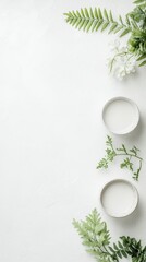 Two white bowls with liquid are arranged with green fern leaves and small white flowers on a white textured background with copy space.