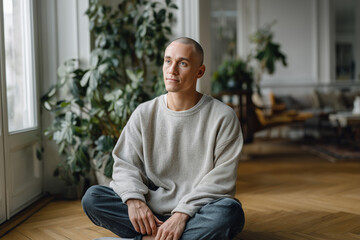 Fototapeta premium Calm young man sitting cross-legged on wooden floor in cozy home interior with plants and natural light