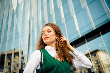 Confident young businesswoman with curly red hair in formal fashion walking outdoors near city...