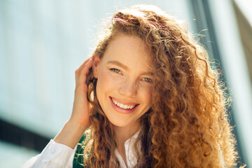 Cheerful young businesswoman with curly hair smiling outdoors on a sunny day near a modern urban...
