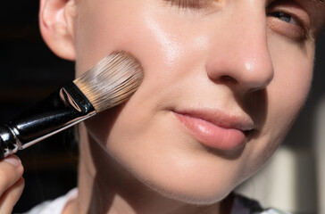 Close-up of a woman applying foundation with brush on cheek. Perfect skin texture, glowing makeup look, natural light. Beauty, skincare, cosmetics, daily facial care scene.