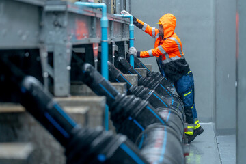 Side view of factory worker or technician with raincoat walk along pipe system and look up to maintenance in area of factory building during raining.