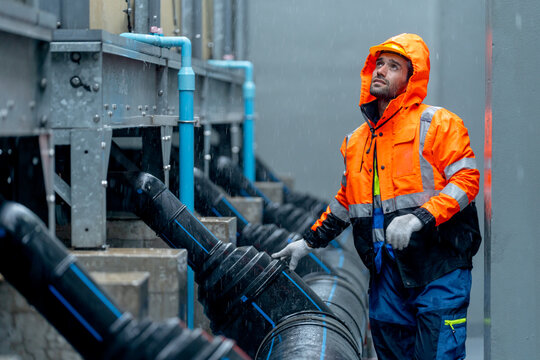 Factory worker or technician with raincoat walk along pipe system and look up to maintenance in area of factory building during raining. - Powered by Adobe