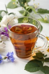Aromatic herbal tea in glass cup, different flowers and green leaves on white wooden table, closeup