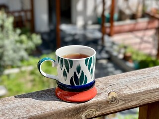Cup of tea in a colorful ceramic mug placed on a wooden railing with a blurred garden and patio background. Cozy outdoor moment, morning tea, relaxation, slow living, casual lifestyle, summer vibes. 