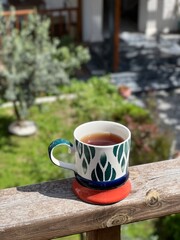 Vertical photo of cup of tea in colorful ceramic mug on wooden railing with blurred garden and patio background. Cozy outdoor scene, morning tea, relaxation, slow living, summer vibes. 