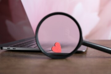 Decorative heart, magnifying glass and laptop on wooden table, closeup. Online dating site