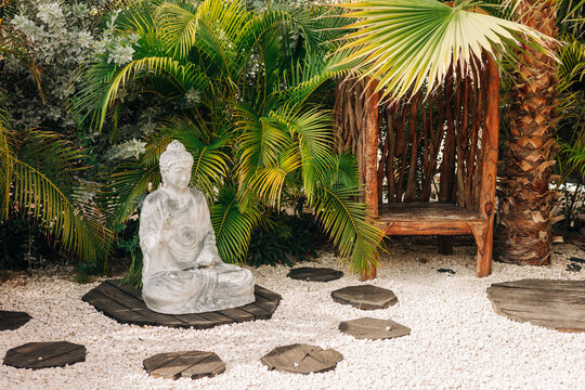 Serene garden setting with stone Buddha statue and wooden chair surrounded by lush greenery - Powered by Adobe