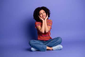 Young stylish woman sitting cross legged against a violet background, wearing casual attire and...