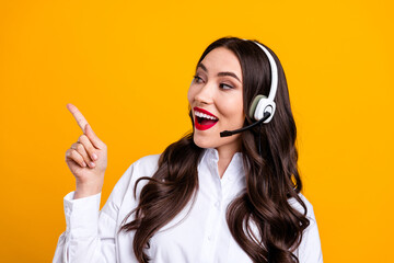 Charming young woman in stylish white shirt with headset pointing with finger on vibrant yellow...