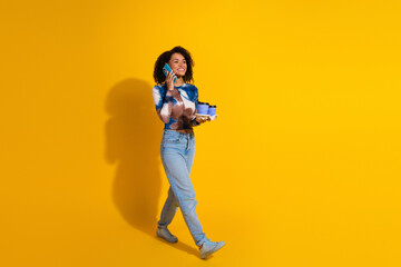 Obraz premium Young woman walking confidently, talking on the phone and holding a gift on a vibrant yellow background