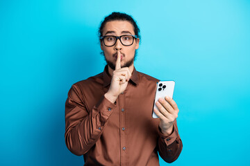 Young businessman holding smartphone making hush gesture against teal background, representing...