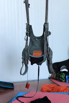 A row of skydiving harnesses and helmets hanging neatly on a metal rack in a well-lit preparation area. The image captures essential parachuting equipment, including protective gear and jumpsuits, rea