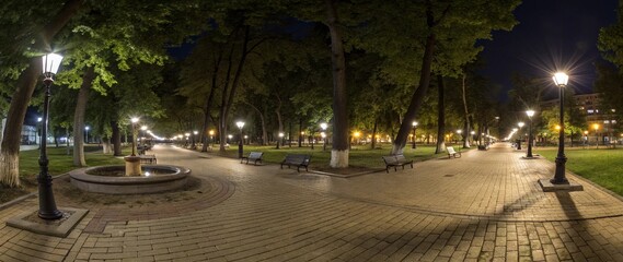 Obraz premium Night urban park with wide stone-paved alley, illuminated by streetlights. Green trees form dense shadows, benches line the walkway, fountain on the left. Buildings visible in the background.