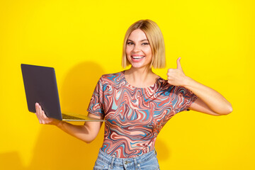 Young stylish blonde woman with a modern laptop, gives a thumbs-up in front of a vibrant yellow...