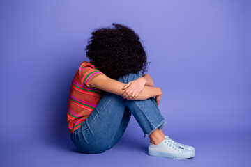 Young woman in casual outfit sitting against purple background, embracing her legs and portraying...