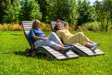 Two beautiful mature women sitting on park benches and talking in city park on sunny summer day	