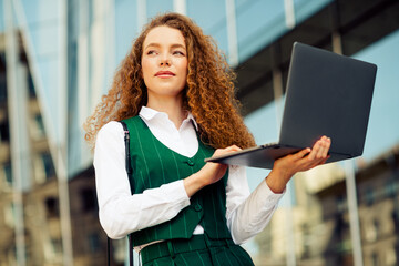Confident young businesswoman with curly hair using laptop outdoors in an urban setting during a...