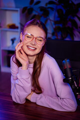 Young female blogger smiling at microphone during live stream in modern home studio with neon...