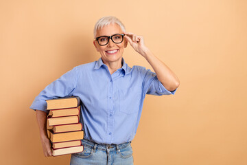 Confident elderly woman in casual attire holding book while adjusting glasses, standing against a...