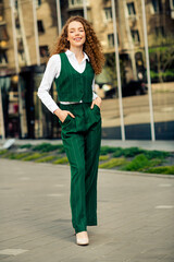 Confident young businesswoman with curly red hair walking in a green waistcoat suit on an urban...