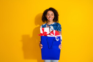 Cheerful young woman holding a blue gift bag with wrapped presents, wearing stylish casual attire...