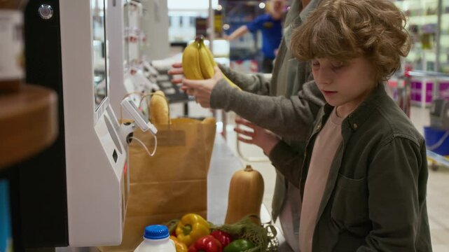 Curly-haired boy scanning bunch of bananas and mesh bag with tomatoes while buying food with mother using self-service checkout in supermarket