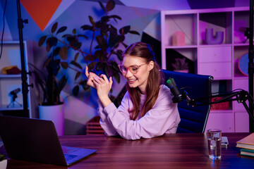Young woman recording a podcast on a microphone in a modern home office studio with vibrant neon...