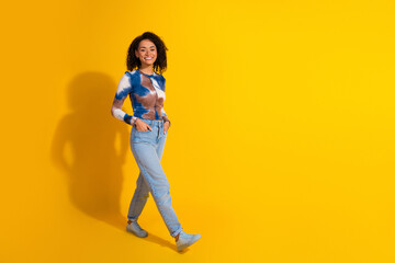 Smiling young woman in casual jeans and trendy tie-dye top walking against a bright yellow...